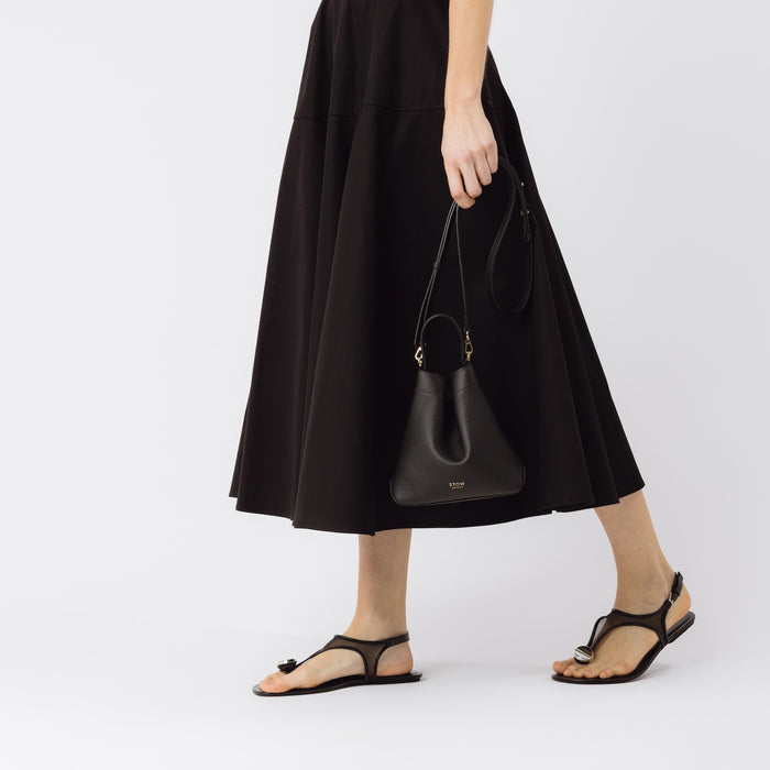 Model holding STOW London's Mini Curve in Black pebbled leather by the detachable leather crossbody strap at her side on a white background