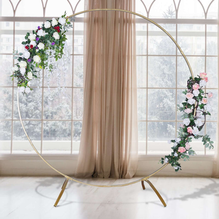 Floral wedding arch with gold frame and greenery against a window backdrop.