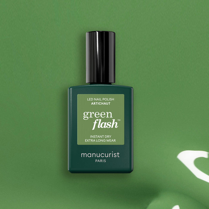 A bottle of Manucurist Artichoke Green Flash LED nail polish sits against a matching green backdrop. The rectangular bottle features a black cap and white text, evoking the lushness of a Moroccan garden.