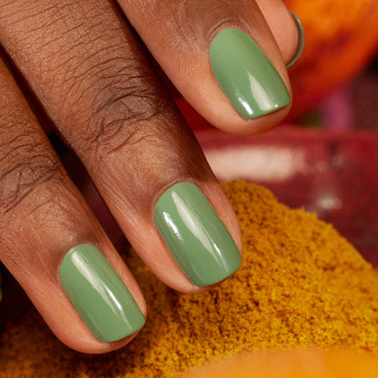 A close-up of a medium brown hand displays neatly manicured nails painted with Manucurist's Artichoke, a glossy herbal green shade. A blurred yellow and red background evokes the feel of a Moroccan garden.