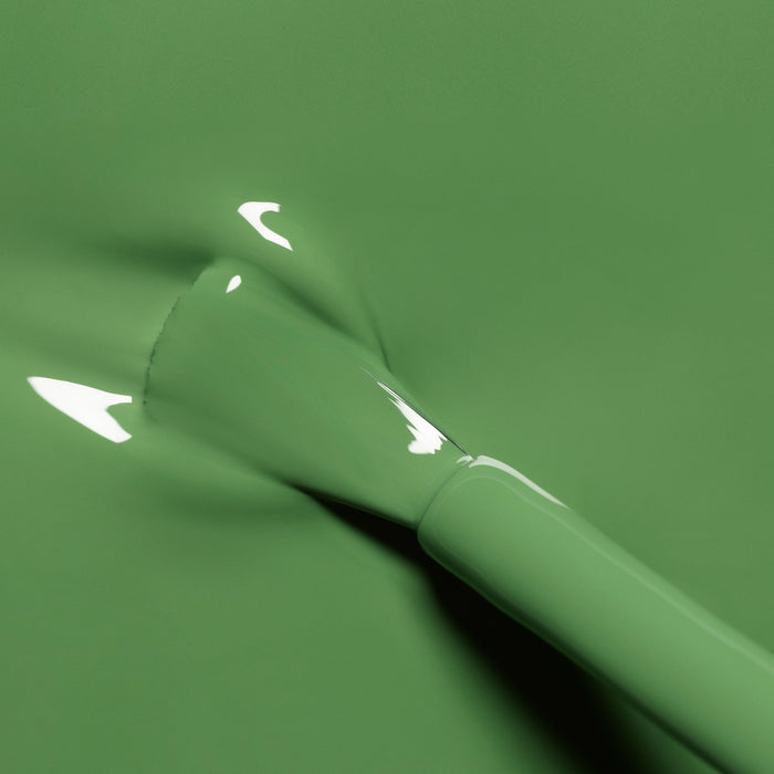 A close-up of a brush applying Manucurist's Artichoke, a glossy herbal green nail polish, onto a smooth surface with light reflecting on the wet color.