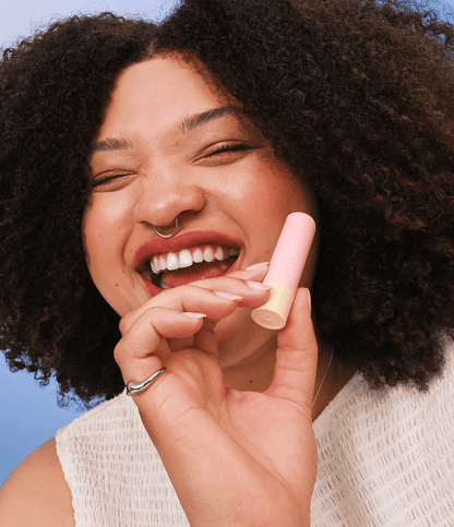 A close-up lifestyle shot of a smiling woman with curly dark hair holding a pink lip balm tube against a bright blue sky background.