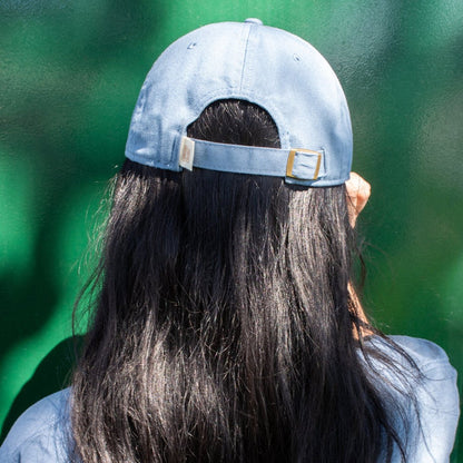back of a woman's head sporting a blue cotton dad hat and blue t-shirt against a green background