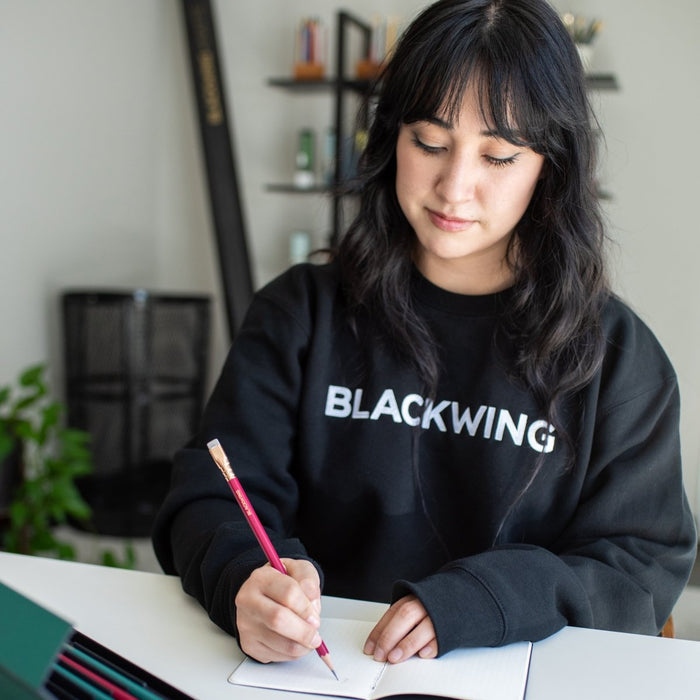 Woman sitting at a desk wearing a black crewneck sweater, writing with a red pencil into a memo book.