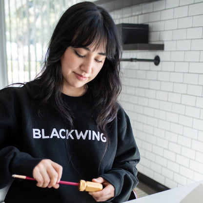 Woman wearing a BLACKWING sweatshirt, sharpening a red pencil with a gold sharpener