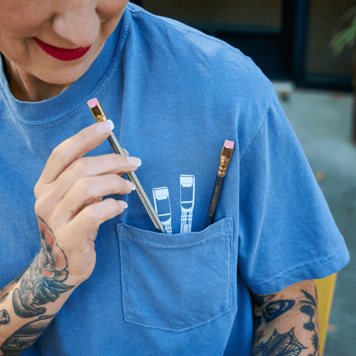Close-up of a woman wearing a blue t-shirt that has white pencil graphics poking out of the pocket. 2 real Blackwing pencils sit in the pocket.