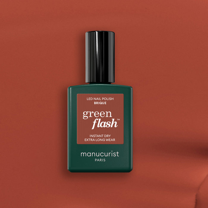 A bottle of Manucurist Brique Green Flash LED nail polish sits on a warm brown background. The dark green bottle with black cap and terracotta-accented label hints at the brick red shade inside.