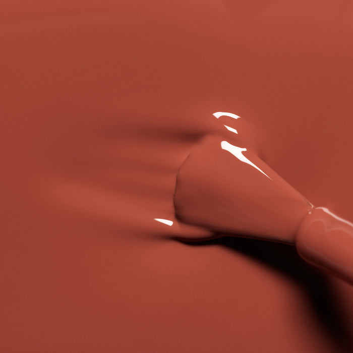 A close-up of a nail polish brush spreading glossy, brick red Manucurist Green Flash Brique nail polish on a smooth surface.