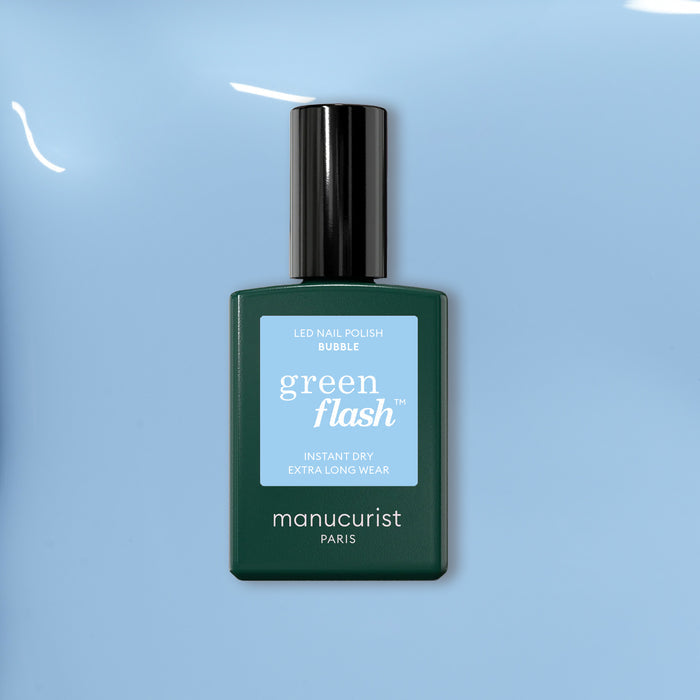 A bottle of Manucurist Bubble nail polish with grey undertones rests on a smooth light blue surface. The dark green bottle features a light blue label and black cap.