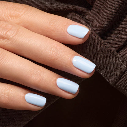 A hand with neatly manicured nails painted in Manucurist’s Bubble, a glossy light blue polish with subtle grey undertones, rests on textured dark brown fabric.
