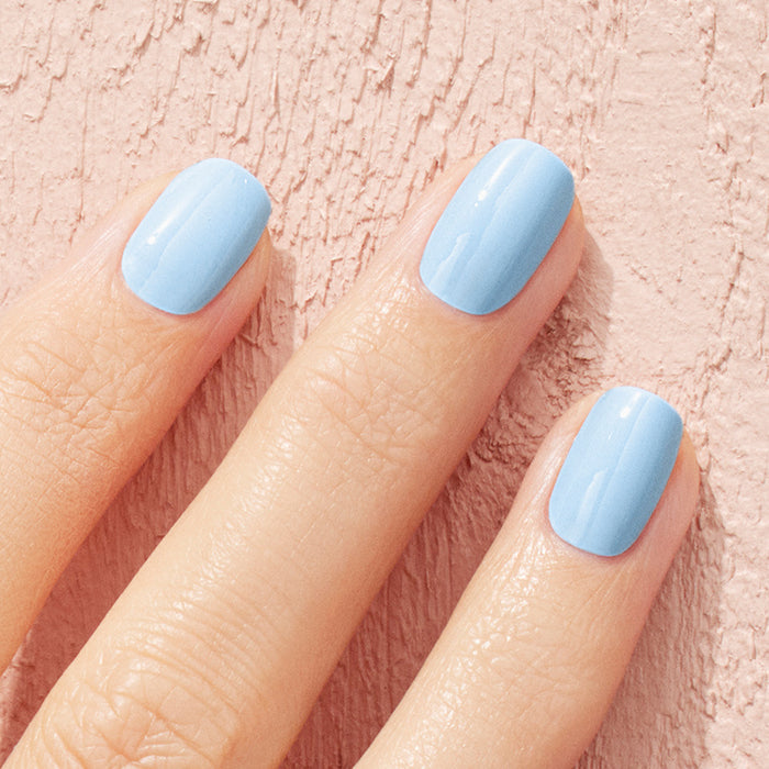 A close-up of a hand with Manucurist's Bubble nail polish—a cool light blue shade with subtle grey undertones—on neatly manicured nails, set against a textured, light pink background.