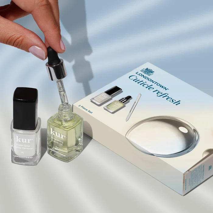 A hand holds a dropper above a LONDONTOWN Cuticle Refresh oil bottle for dry cuticles, placed next to nail polish and the LONDONTOWN Cuticle Refresh kit box on a light surface.
