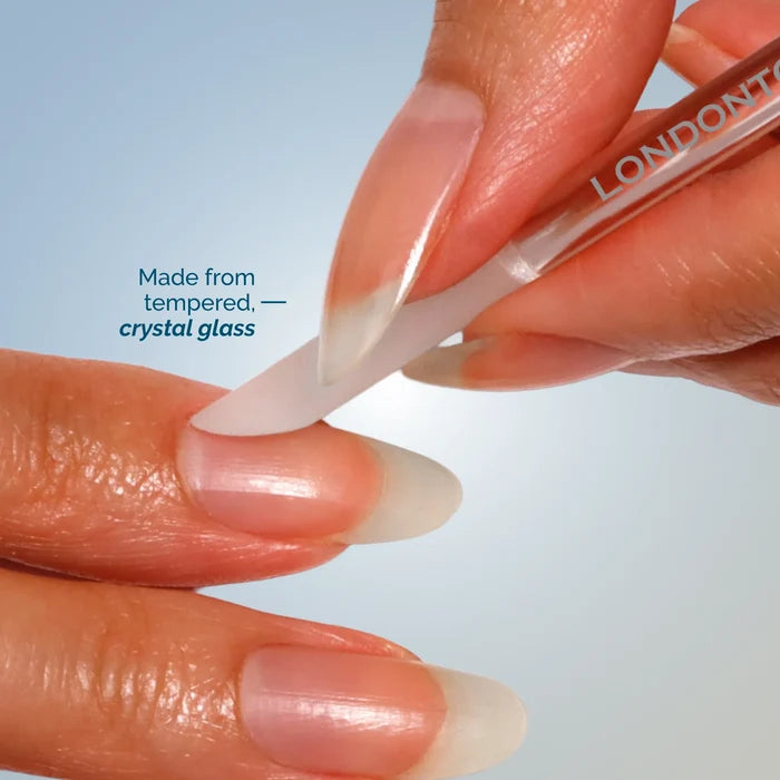 A hand uses LONDONTOWN's Cuticle Refresh glass nail file to shape natural fingernails against a light blue background, highlighting its tempered crystal glass—ideal for gentle filing and cuticle care.