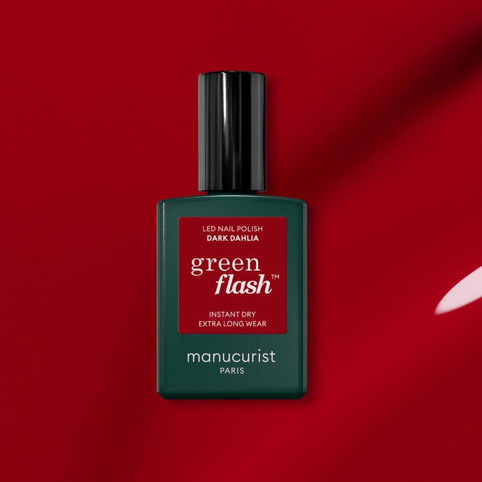 A bottle of Manucurist Dark Dahlia gel polish sits on a red background. The green bottle with a black cap and red label showcases the rich hue of the Dark Dahlia shade.