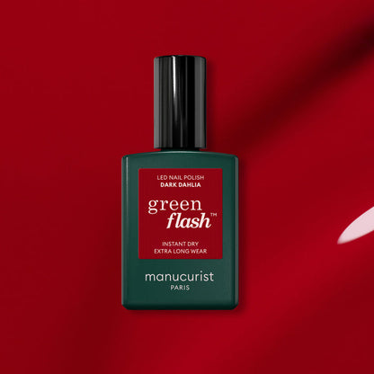 A bottle of Manucurist Dark Dahlia gel polish sits on a red background. The green bottle with a black cap and red label showcases the rich hue of the Dark Dahlia shade.