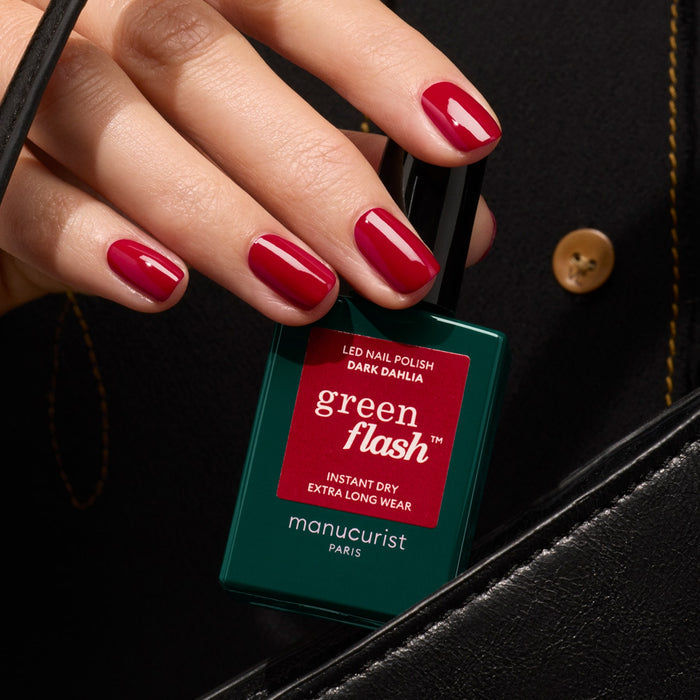 A hand with glossy red nails holds a bottle of Manucurist Dark Dahlia gel polish near a black handbag with a gold button.