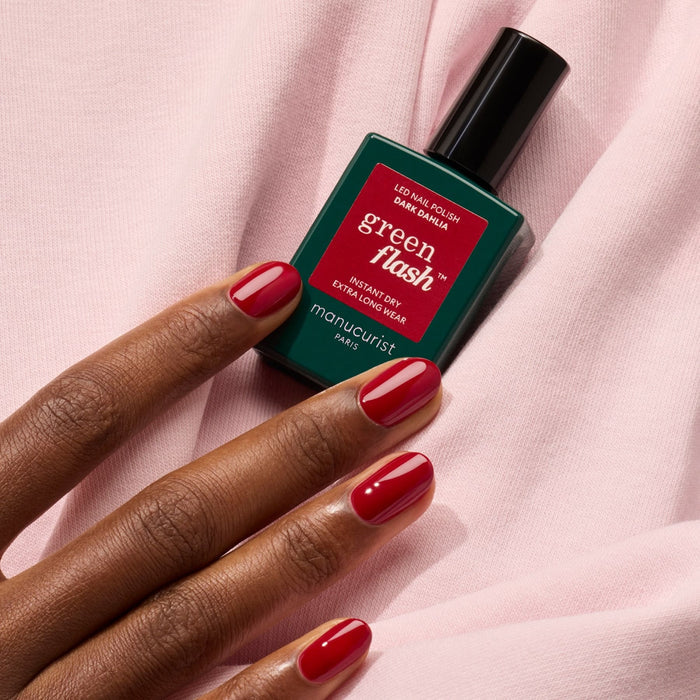 A hand with neatly manicured red nails holds a bottle of Manucurist Dark Dahlia gel polish against a soft pink fabric background.