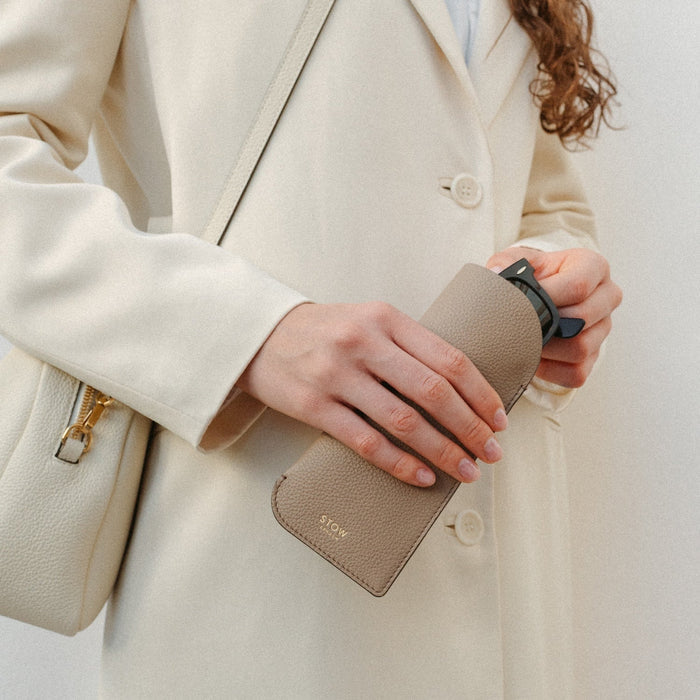 Model holding STOW London's Glasses Case in Taupe pebbled leather, featuring a gold logo