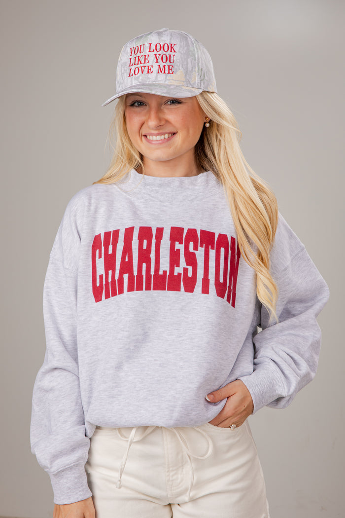 Woman wearing a gray sweatshirt with 'Charleston' printed on it, a white cap, and light pants against a neutral background.