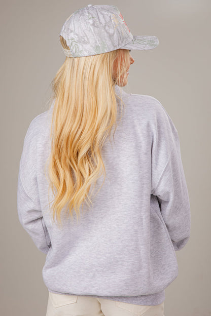 Person wearing a gray mocneck sweatshirt and cap with long blonde hair on a beige background