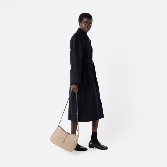 Model holding STOW London's Beth bag by the detachable crossbody strap in Warm Taupe leather at her side featuring gold stainless steel hardware on a white background