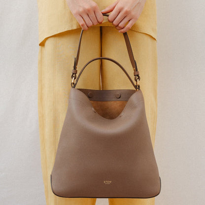 Model holding STOW London's Curve in Mocha leather featuring the detachable shoulder strap and top handle with gold hardware and details