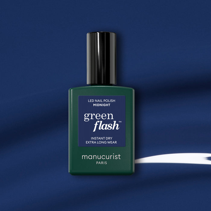 A bottle of Manucurist Green Flash gel polish in the shade Midnight rests on a dark blue background with a white curved streak, featuring a dark green bottle, black cap, and navy label that highlights the midnight blue polish.