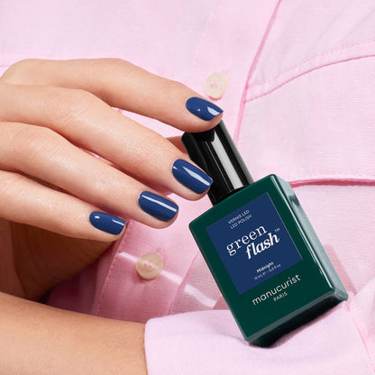 A hand with glossy dark blue nails holds a bottle of Manucurist Green Flash gel polish in the shade Midnight against a light pink shirt background.