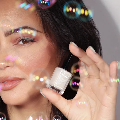 A person holds a bottle of LONDONTOWN Milk Bath demi-sheer cream nail color near their face, surrounded by shimmering bubbles with soap bubble iridescence.