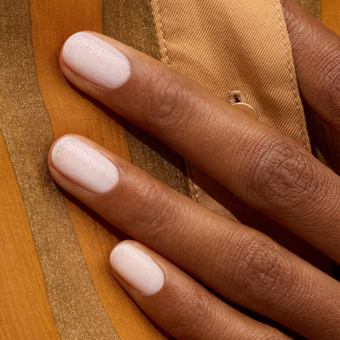 A close-up of a hand with dark skin and short, neat nails polished with Manucurist's Nebula holographic gel, resting on gold and brown striped fabric.
