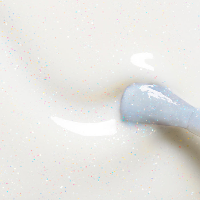 Close-up of Manucurist’s Nebula nail polish brush applying milky white polish with multicolored glitter on a smooth surface, revealing a glossy, holographic-like finish with scattered sparkling specks.