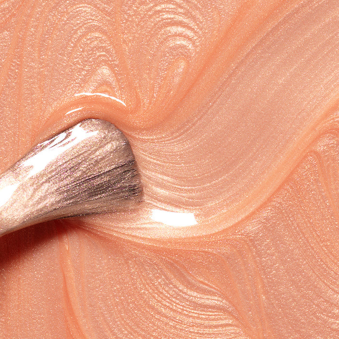 A close-up of a brush gliding through Manucurist’s Pamplemousse nail polish, creating smooth, glossy waves in peachy shimmer with a metallic sheen.