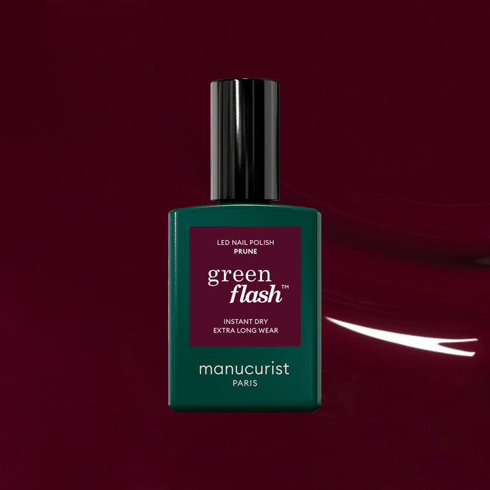 A bottle of Manucurist Prune Green Flash LED nail polish, with a green base and dark cap, stands out against a deep burgundy background, highlighting the elegance of its rich wine red shade.