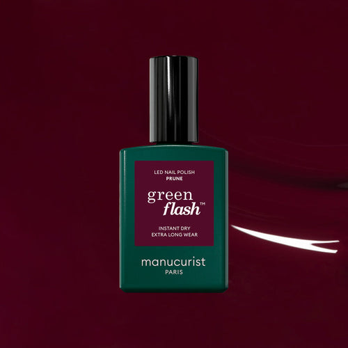 A bottle of Manucurist Prune Green Flash LED nail polish, with a green base and dark cap, stands out against a deep burgundy background, highlighting the elegance of its rich wine red shade.