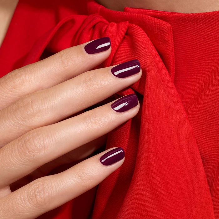 A hand with neatly manicured nails painted in Prune, a glossy vintage purple gel polish by Manucurist, rests on bright red fabric with a tied bow.