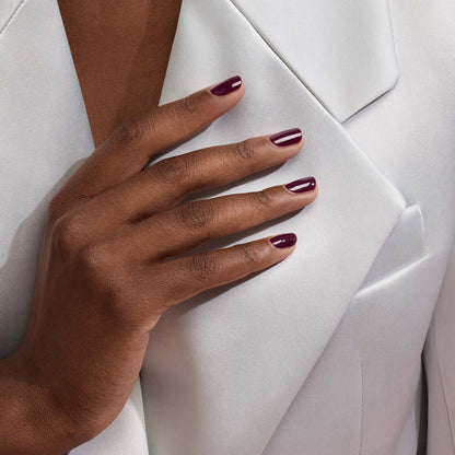 A close-up shows a hand with Manucurist’s Prune gel polish, a deep wine red, resting on a light blazer lapel—highlighting well-groomed nails and smooth fabric.