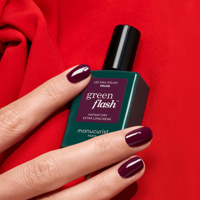 A hand with deep wine red gel polish (Prune by Manucurist) holds a green bottle of Green Flash LED nail polish against a bright red fabric background.