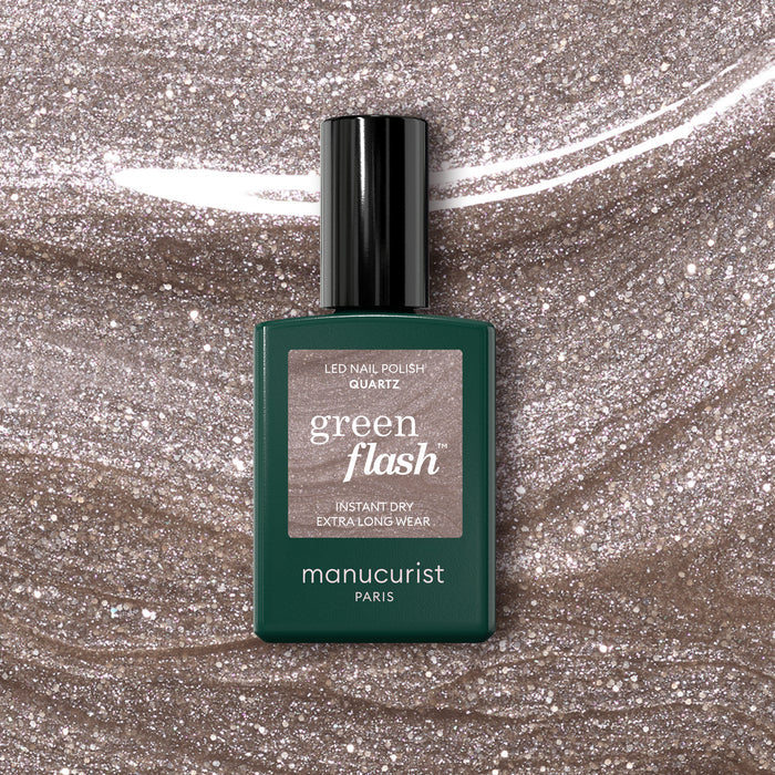 A bottle of Manucurist Quartz Green Flash semi-permanent nail polish sits on a glittery beige surface that matches the soft, refined pinkish-beige shade shown on the label.