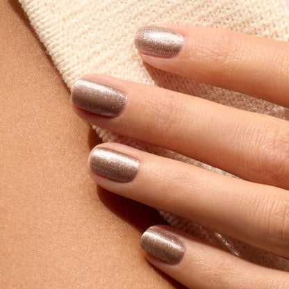 A close-up of a hand with short, neatly manicured nails painted in Manucurist's Quartz glittery brown polish, resting on beige textured fabric and bare skin.