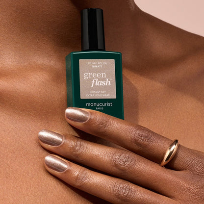 A hand with chestnut brown, semi-permanent polished nails holds a green bottle of Manucurist Quartz nail polish. The person wears a simple gold ring, and the background is soft beige.
