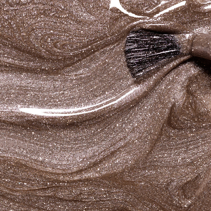 A close-up of a brush swirling through Manucurist's Quartz, a shimmery, metallic bronze liquid makeup that creates glossy, marbled patterns with a chestnut brown finish.