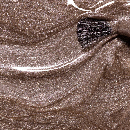 A close-up of a brush swirling through Manucurist's Quartz, a shimmery, metallic bronze liquid makeup that creates glossy, marbled patterns with a chestnut brown finish.