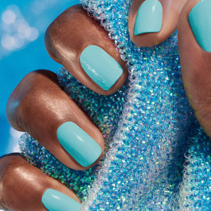 A hand with medium brown skin tone showcases neatly manicured nails painted in Manucurist’s energizing Riviera blue nail polish, holding a sparkling blue textured fabric with iridescent beads against a matching blue background.