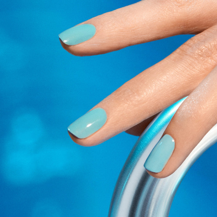 A close-up of a hand with Riviera gel polish by Manucurist on the nails, resting on a shiny metal pool ladder over clear blue water.