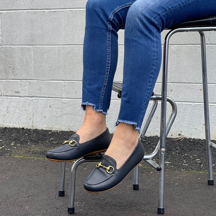Person wearing Tango's navy loafers with gold buckles sitting on a metal stool against a gray wall. Comfortable, quality footwear.